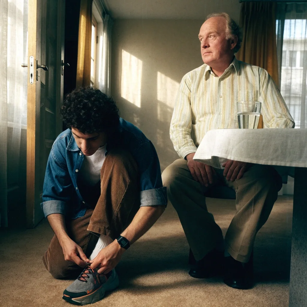 A younger man tying a shoe and an older man seated at a stone table with water and a dish of capsules